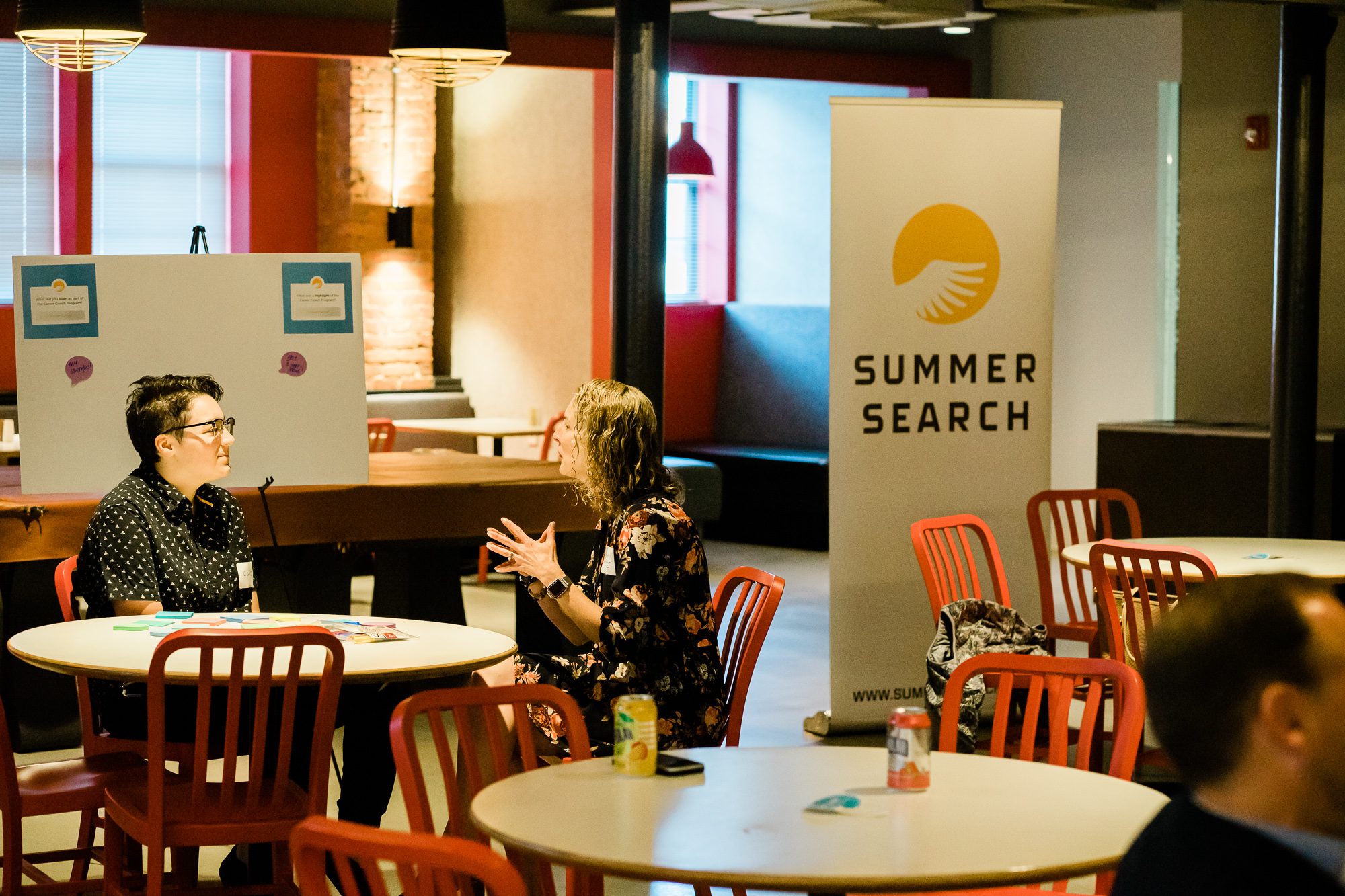 Two people at a table with a Summer Search banner behind them